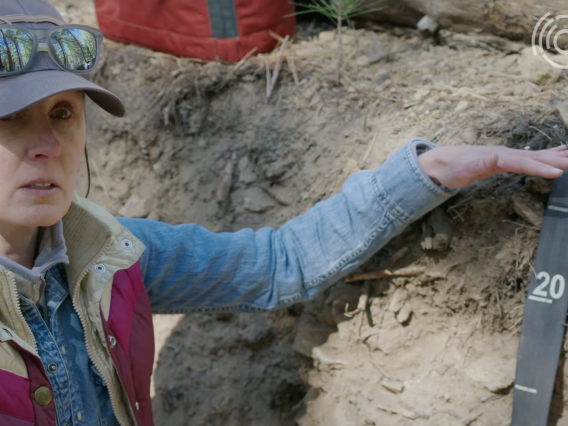 A woman in a baseball hat and sunglasses kneels in a soil pit, explaining layers of soil.