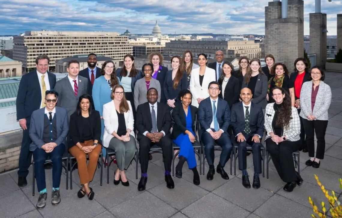 A group of people in professional attire smile at the camera with white brick buildings in the background.