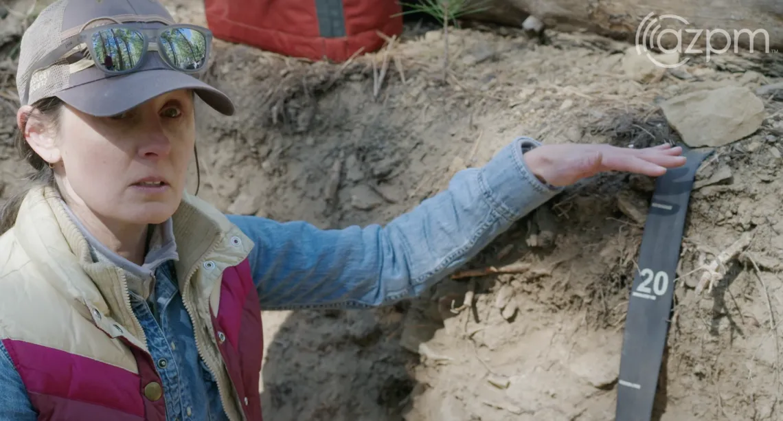 A woman in a baseball hat and sunglasses kneels in a soil pit, explaining layers of soil.