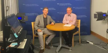 Two men sit at a round table in a recording studio with a blue backboard.