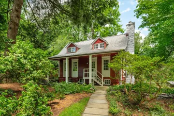 A quaint red house with white doors and windows and chimney stands in a green wood under a blue sky.