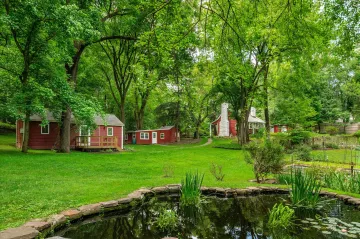 A pond with reeds; and behind, red buildings with white trim under a forest on a green, manicured landscape.